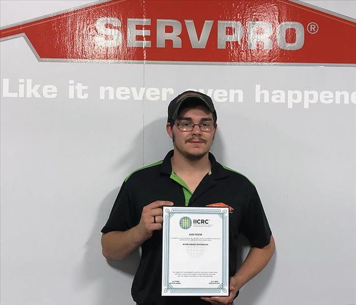 Alex Foster stands in front of a wall featuring the SERVPRO logo.  He holds a framed IICRC WRT certification.